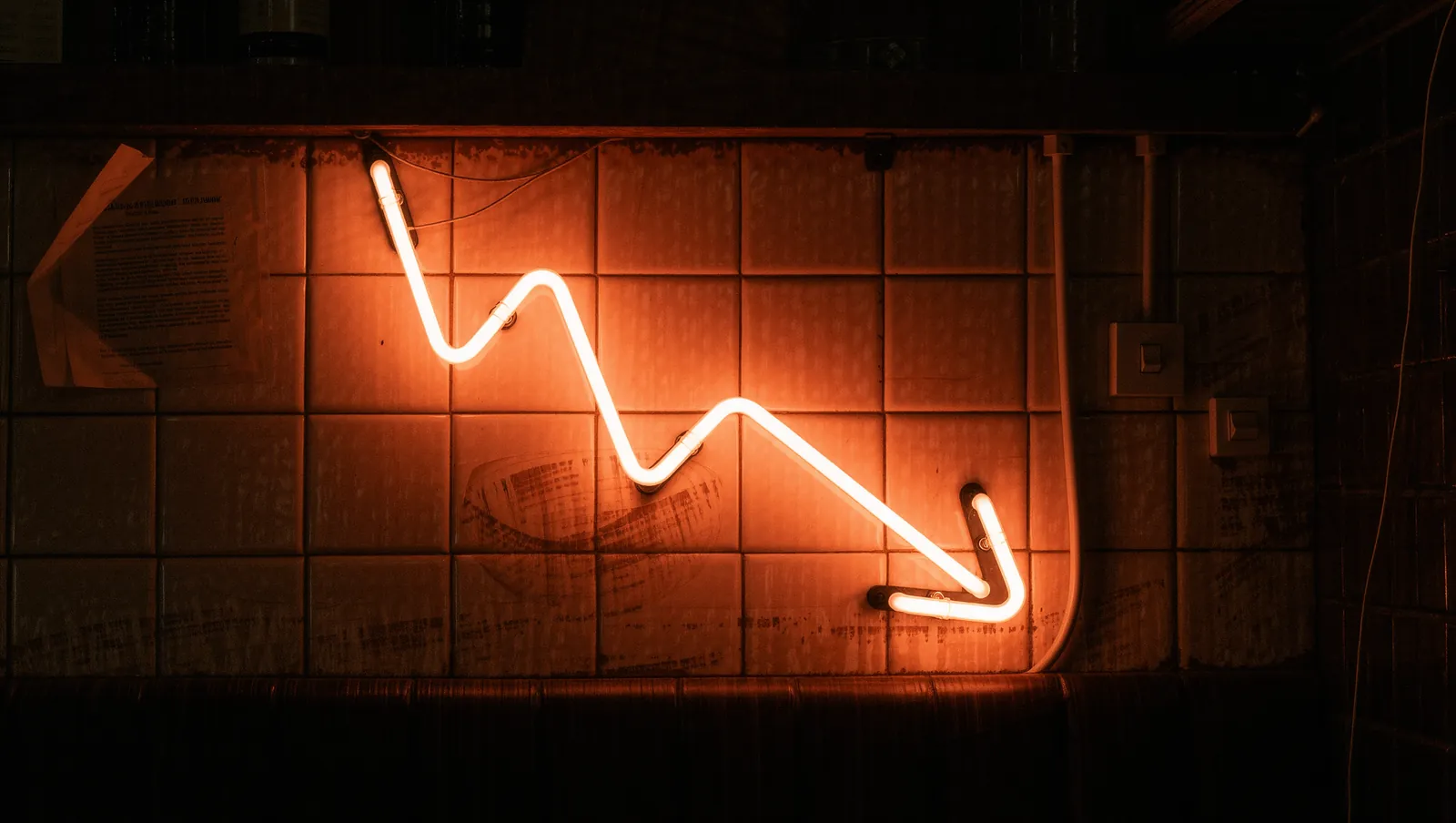A neon sign shaped like a falling price chart, mounted on the tiled wall of a late-night bar